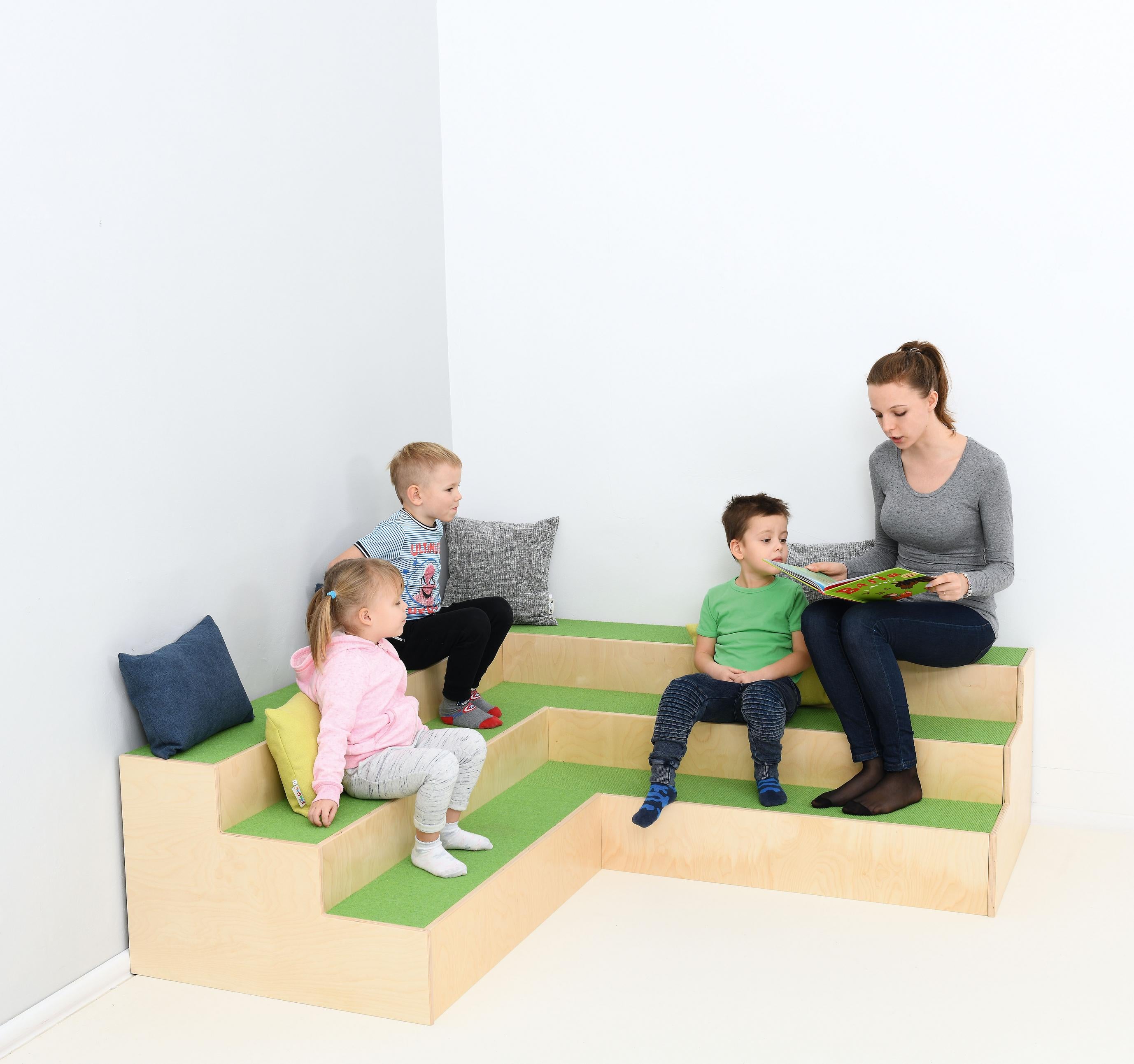 Kinder sitzen auf Stufenpodest, genießen Vorlesestunde.