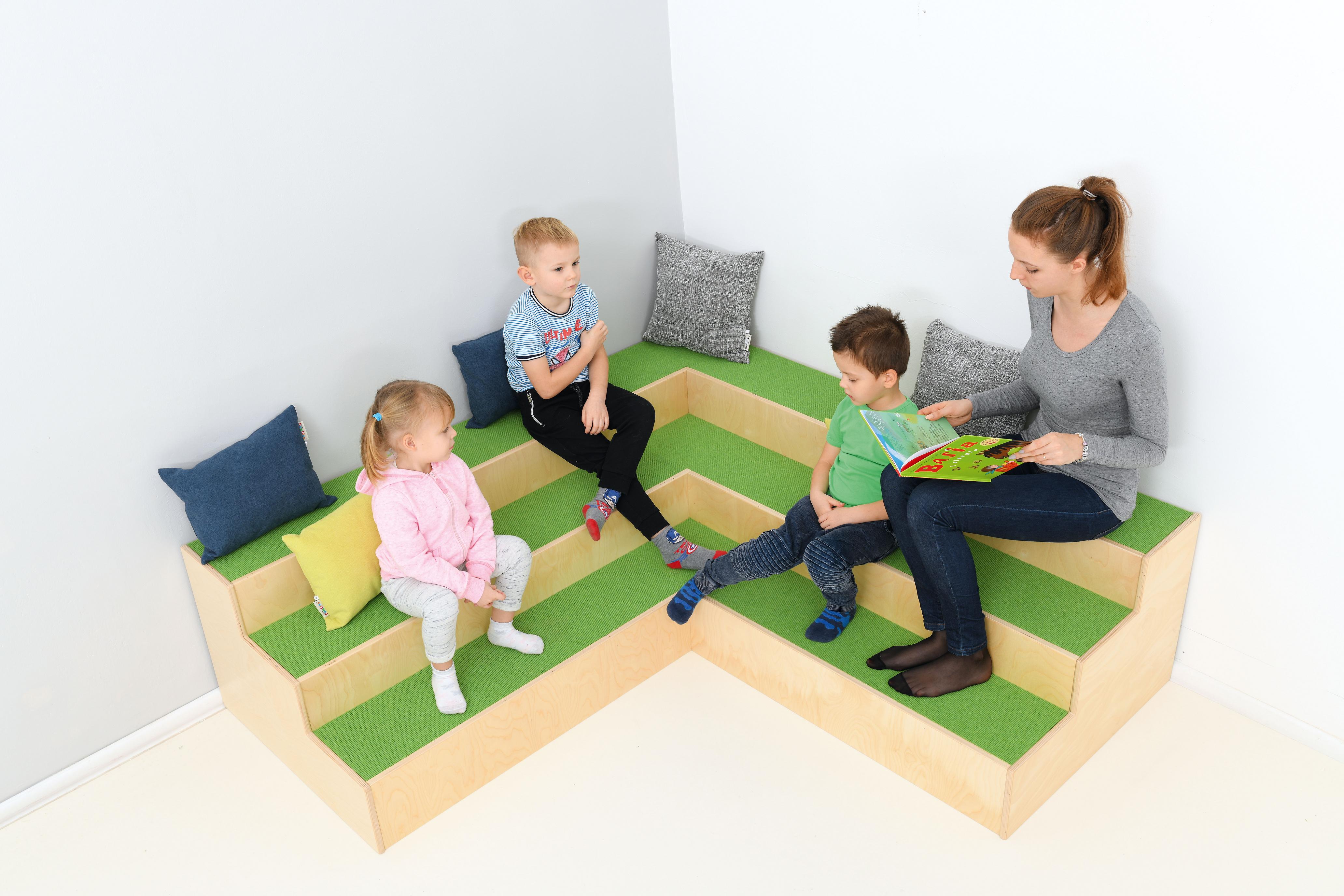 Kinder und Betreuerin auf Stufenpodest-Sitzecke beim Vorlesen.