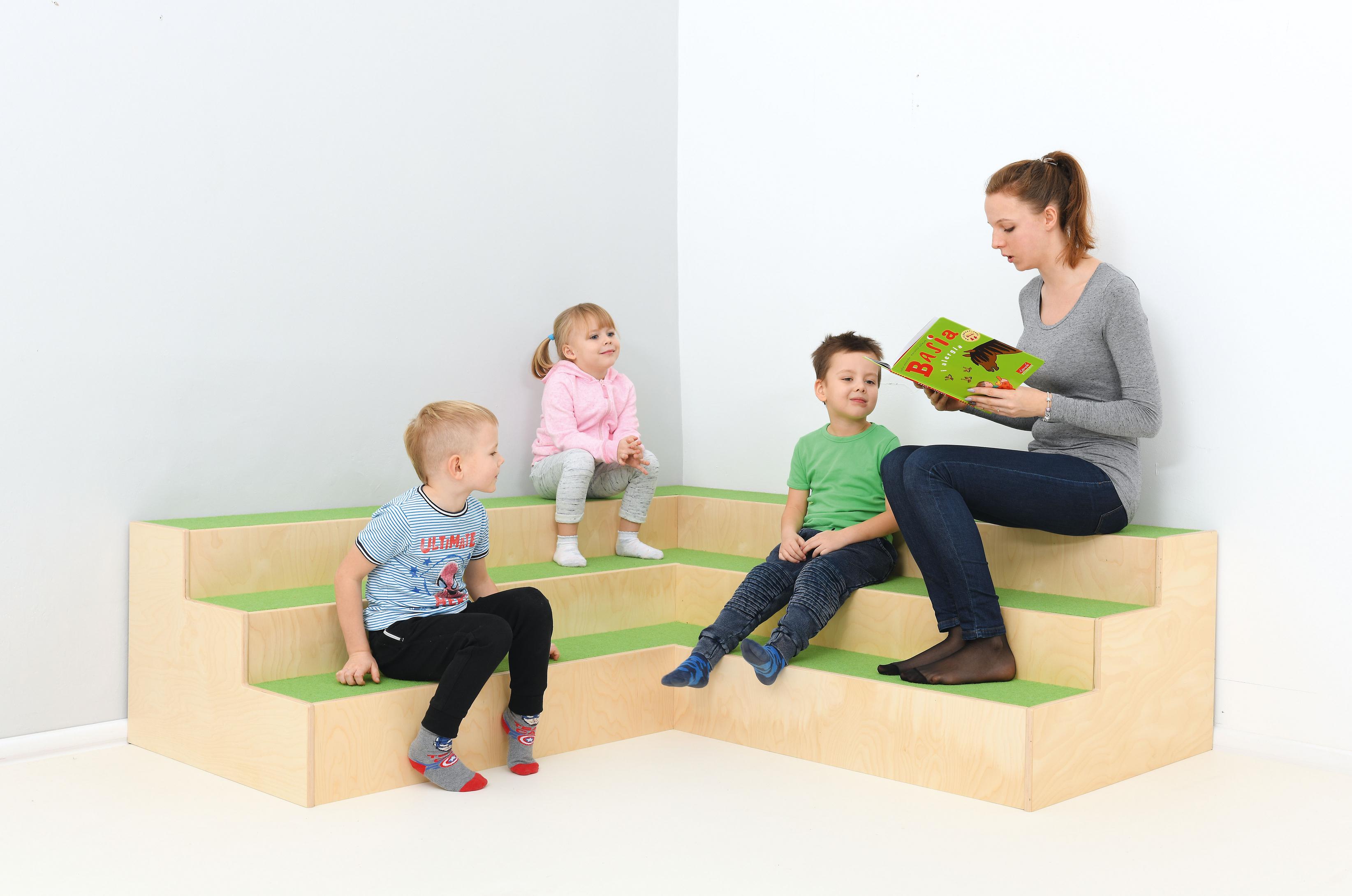 Kinder sitzen auf Stufenpodest, Erwachsene liest Buch vor.